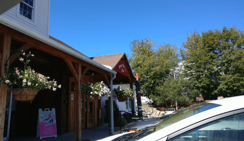A sunny day at a building with wooden architecture, featuring hanging flower baskets and a sign. Cars are parked nearby.