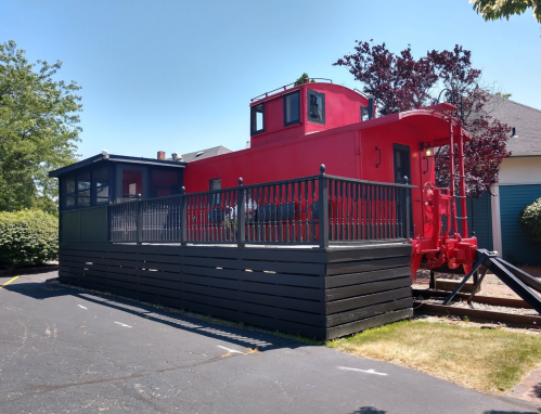 A bright red caboose converted into a building, surrounded by a black wooden deck and greenery.
