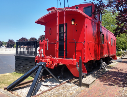 A bright red caboose on a track, surrounded by greenery and a clear blue sky.