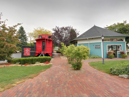 A red caboose next to a green building, surrounded by trees and a brick pathway. Overcast sky above.