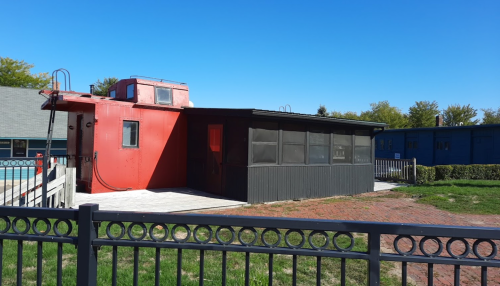A red building with a flat roof and screened porch, surrounded by greenery and a fence, under a clear blue sky.