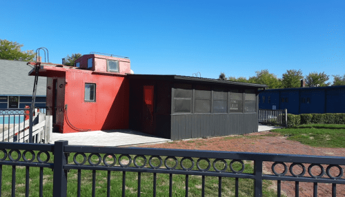 A red building with a flat roof and screened porch, surrounded by greenery and a fence, under a clear blue sky.