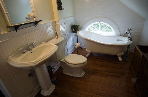 A vintage bathroom featuring a clawfoot tub, pedestal sink, and toilet, with wooden flooring and a round window.