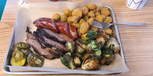 A tray of barbecue featuring brisket, sausage, fried okra, Brussels sprouts, and pickled jalapeños and cucumbers.