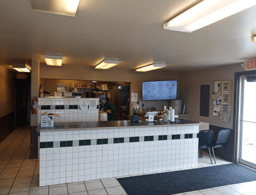 Interior of a casual eatery with a counter, menu board, and seating area. Staff member visible behind the counter.