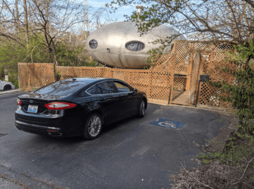 A black car parked in front of a unique, futuristic silver structure surrounded by trees and a wooden fence.