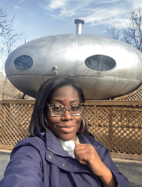 A person with glasses poses in front of a futuristic, silver spaceship-like structure on a sunny day.