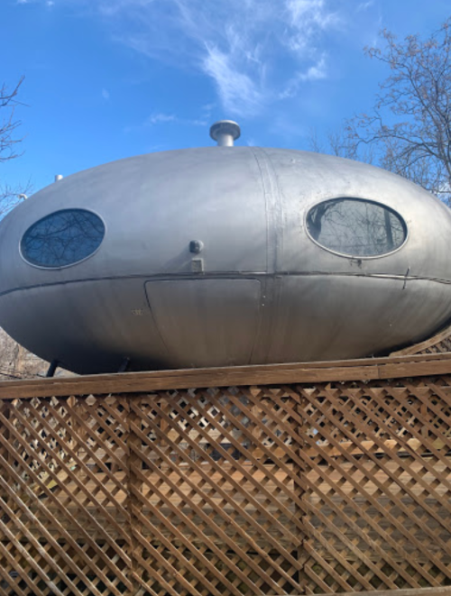 A metallic, oval-shaped structure with round windows, perched on a wooden deck against a blue sky.