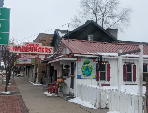 A quaint hamburger restaurant with a red sign reading "Open Hamburgers," surrounded by snow and a white picket fence.