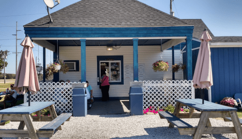A small blue and white building with a porch, picnic tables, and a person ordering at a window. Flowers adorn the area.