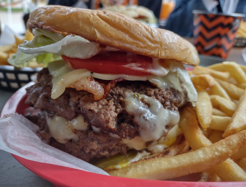 A large, stacked burger with lettuce, tomato, bacon, and melted cheese, served with a side of fries in a red basket.