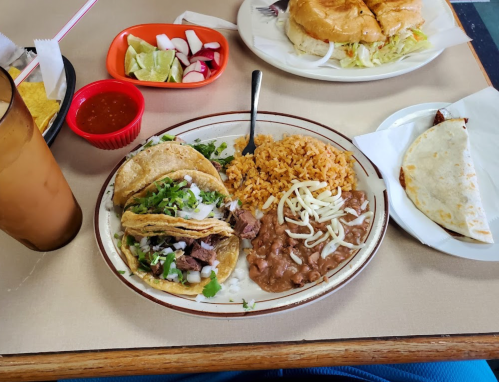 A plate with three tacos topped with onions and cilantro, rice, refried beans, and a side of lime and radishes.