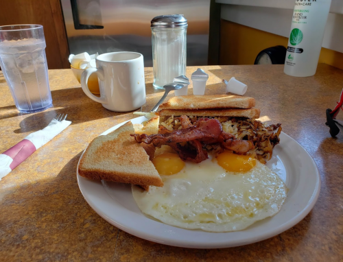 A plate of breakfast with two eggs, bacon, hash browns, and toast, alongside a glass of water and a coffee cup.
