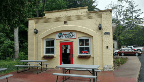 A small, yellow building with a red door and "Open" sign, featuring outdoor seating and surrounded by greenery.