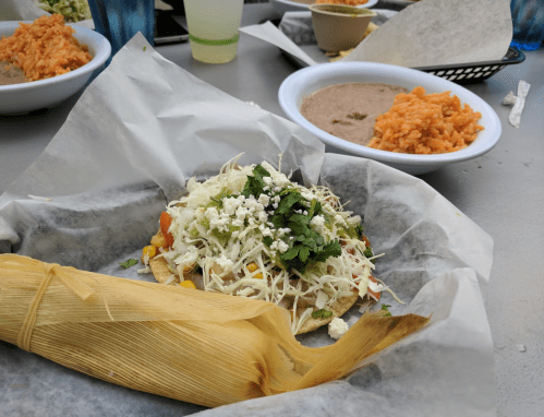 A tamale topped with vegetables and cheese, served with rice and beans in the background.