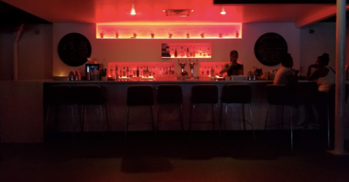 A dimly lit bar with red lighting, showcasing a long counter and patrons seated on high stools.