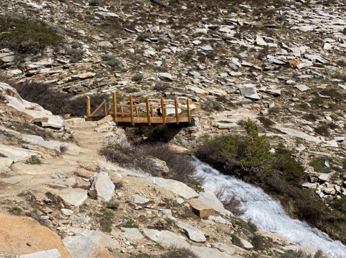 A wooden bridge spans a rocky landscape with a small stream flowing nearby, surrounded by sparse vegetation.