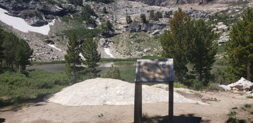 A scenic view of a rocky landscape with a lake, featuring a signpost in the foreground and trees surrounding the area.