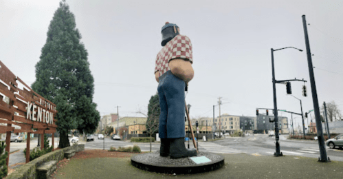 A large statue of a lumberjack stands at a street corner in Kenton, with a sign welcoming visitors to the area.
