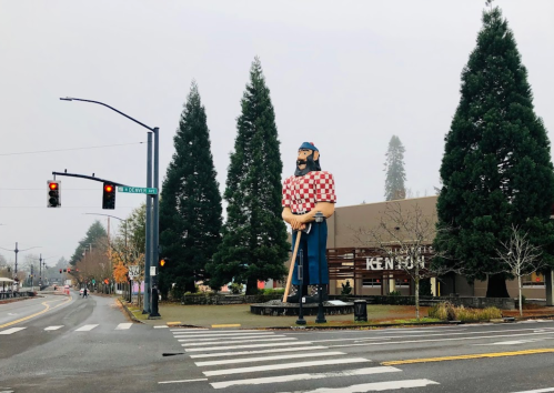 A large statue of a lumberjack stands near a street intersection, surrounded by tall trees and a foggy sky.