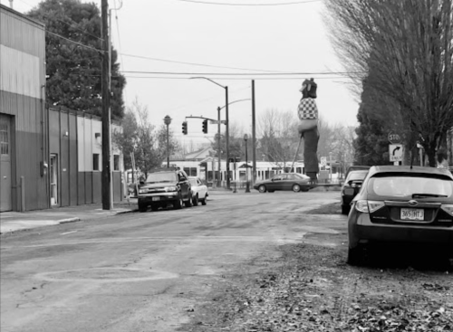 A black and white street scene featuring parked cars and a large statue in the background. Trees line the street.