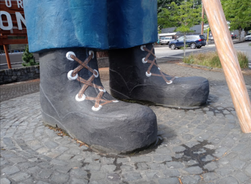 Giant statue boots on a cobblestone surface, with a wooden stick nearby and greenery in the background.