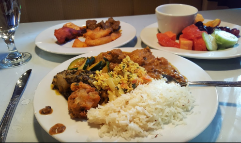 A plate of rice with mixed vegetables, curry, and shrimp, alongside plates of fried chicken and fresh fruit.