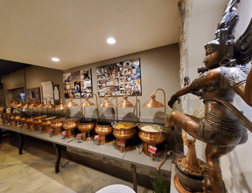 A buffet with copper pots filled with food, alongside a statue of a dancing figure and a wall of photos.