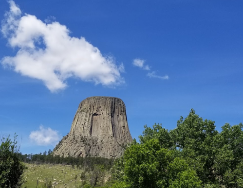A tall, flat-topped rock formation under a blue sky with a few clouds, surrounded by green trees.