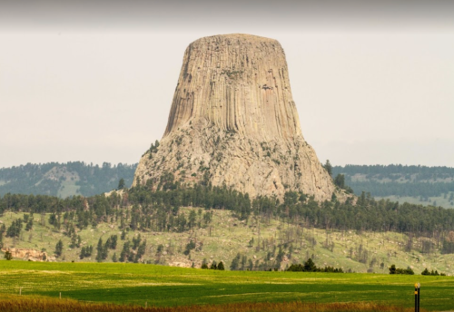 A towering rock formation rises above green fields and trees under a cloudy sky.