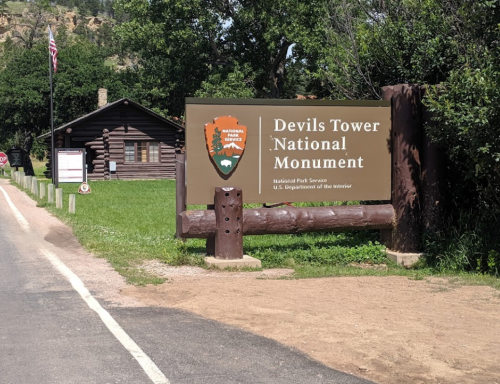 Sign for Devils Tower National Monument, with a rustic building and trees in the background. Roadway visible in front.