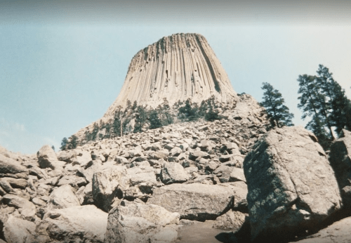 A towering rock formation rises above a rocky landscape, surrounded by sparse trees under a clear blue sky.