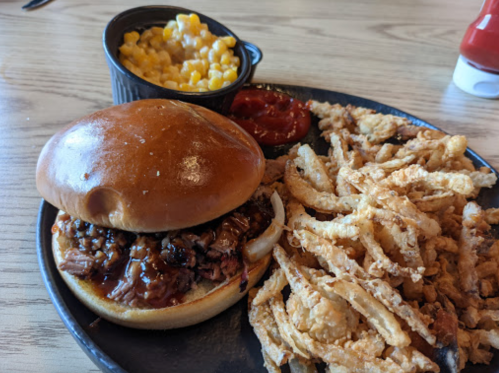 A plate with a BBQ sandwich, crispy onion rings, mac and cheese, and ketchup on a wooden table.