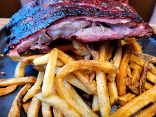 A plate of crispy French fries topped with a large, smoky rib with a charred crust.