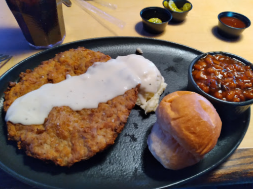 A plate with fried chicken, creamy gravy, mashed potatoes, baked beans, and a soft roll, with a drink in the background.