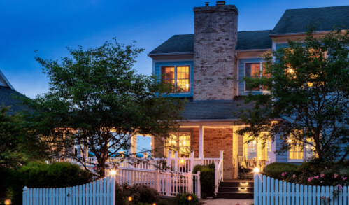 A charming two-story house at dusk, with warm lights glowing from the windows and a white picket fence.