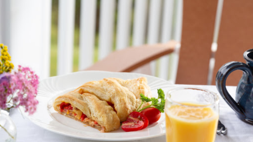 A plate with a flaky pastry filled with tomatoes, alongside a glass of juice and a coffee pot, set on a table.