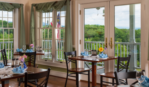 A cozy restaurant with wooden tables set for dining, featuring large windows and a view of greenery outside.
