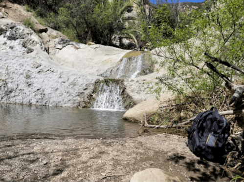 A serene landscape featuring a small waterfall and pool, with a backpack hanging from a tree branch nearby.