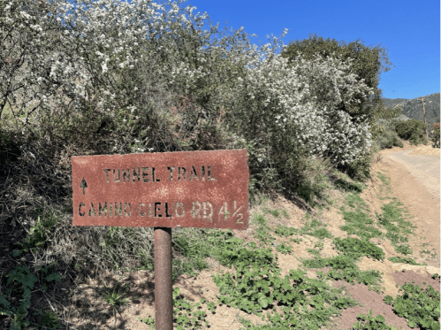 A weathered sign points to "Tunnel Trail" and "Camino Cielo Rd 4½," surrounded by greenery and blue sky.