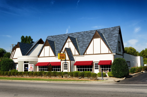 A charming café with a Tudor-style design, featuring a sloped roof and red awnings, surrounded by neatly trimmed hedges.