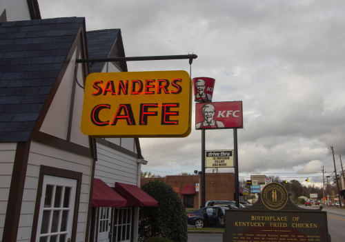 Sign for Sanders Cafe with a KFC sign in the background, featuring a historical marker for the birthplace of Kentucky Fried Chicken.