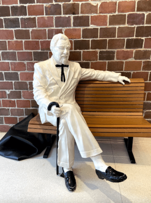 A white statue of a man in a suit and bow tie, sitting on a bench with a cane, against a brick wall.
