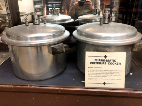 Two vintage Mirro-Matic pressure cookers displayed in a glass case, with a descriptive plaque nearby.