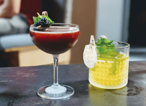 Two cocktails on a table: a red drink in a coupe glass and a yellow drink in a textured glass, both garnished with herbs.