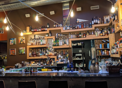 A bar with a wooden shelf displaying various bottles of liquor, illuminated by hanging lights and colorful artwork on the walls.