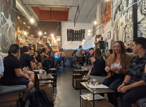 A lively bar scene with patrons seated at tables, enjoying drinks and conversation in a stylish, artistic setting.