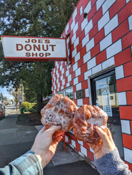 Two hands hold glazed donuts in front of Joe's Donut Shop, featuring a red and white checkered exterior.
