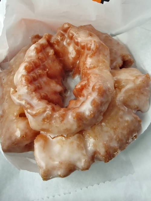 A close-up of a glazed, cruller-style donut with a light brown color, resting on white paper.