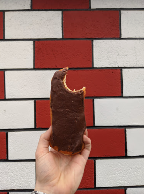 A hand holds a chocolate-covered pastry with a bite taken out, against a red and white brick wall.